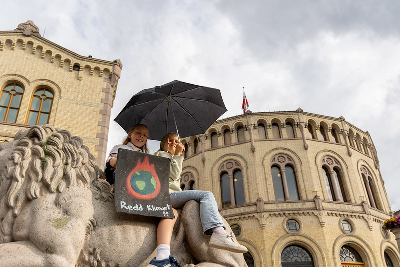 To barn sitter med plakat foran Stortinget under markeringen "Stå opp" for klima og natur før stortingsvalget 2025.