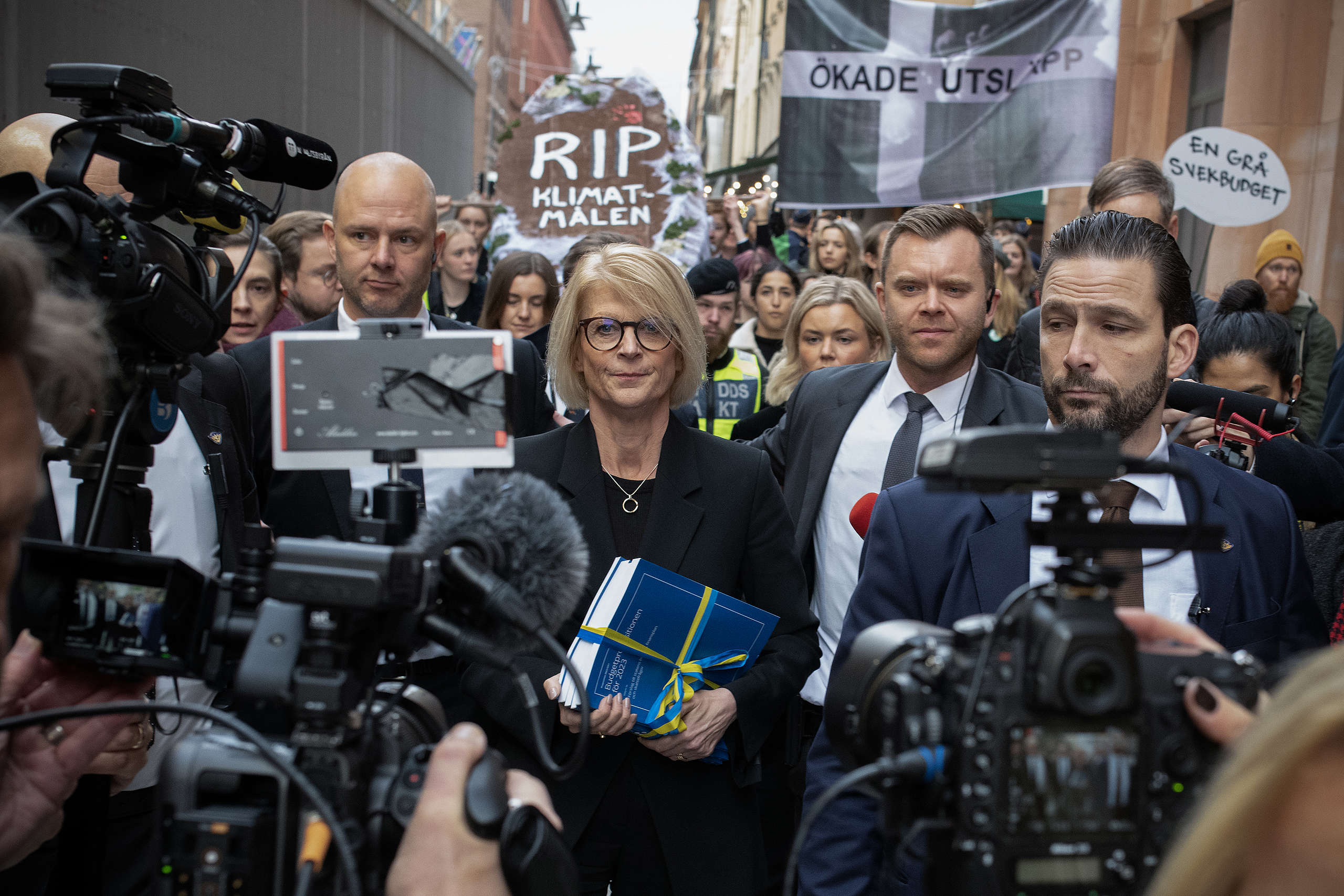 Representanter från regeringen, däribland finansminister Elisabeth Svantesson (M), går den klassiska budgetpromenaden utanför riksdagen i Stockholm. I bakgrunden syns Greenpeace skyltar som lyder "RIP klimatmålen" och "Ökade utsläpp".