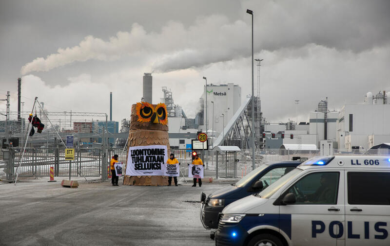 Polisen står framför aktivisterna från Greenpeace som blockerar ingången till pappersmassafabriken. En enorm uggla står vid aktivisterna, med massafabriken bakom. 