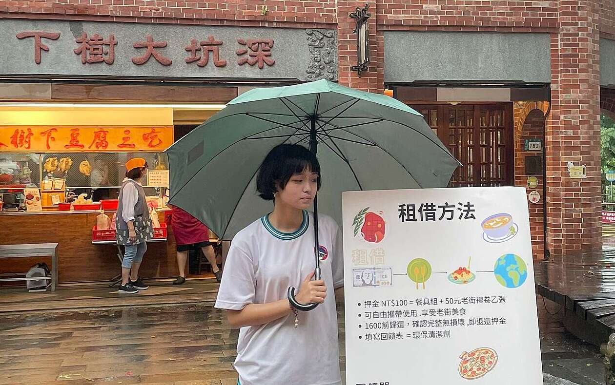 學生戴謹昕在深坑老街的大樹入口處,向遊客宣傳餐具租借方法。