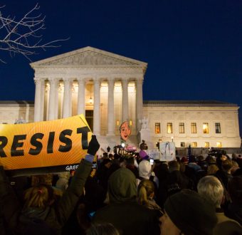 A photo showing a crowd of people protesting at night outside of the US Supreme court. In the foreground, there is a person holding a sign that reads “RESIST.”